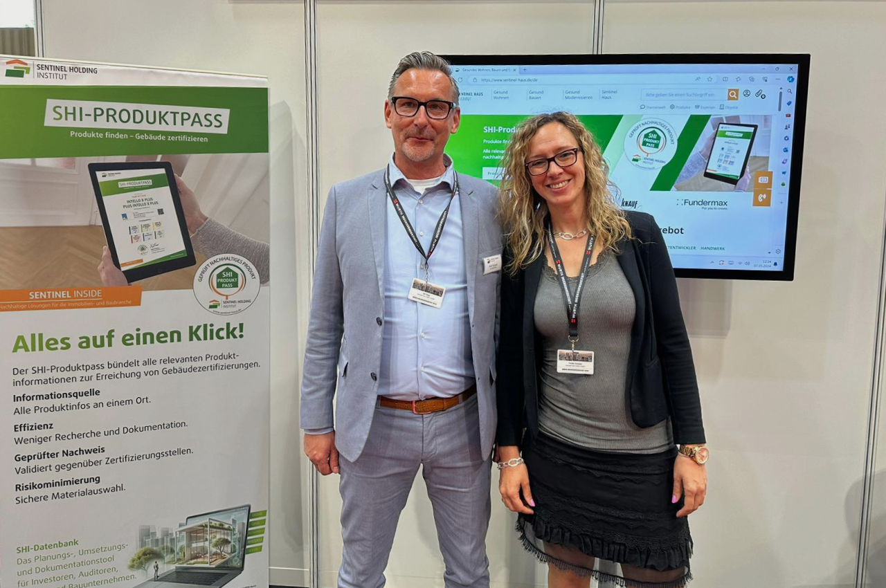 Das Bild zeigt zwei Personen, einen Mann und eine Frau, die vor einem Messestand stehen. Beide tragen Namensschilder und lächeln in die Kamera. Der Mann trägt einen hellblauen Anzug mit weißem Hemd und Krawatte, während die Frau eine schwarze Jacke über einem grauen Kleid trägt. Im Hintergrund ist ein großes Poster mit der Aufschrift "SHI-PRODUKTPASS" und weiteren Informationen sowie ein Monitor mit einer Präsentation zu sehen. Der Stand wirkt professionell und informativ gestaltet, und die beiden Personen sind das Standpersonal, das für Fragen und Informationen zur Verfügung steht.