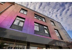 Fassade mit irisierenden Paneelen in Violett- und Pinktönen sowie großen Fenstern, vor einem teils bewölkten Himmel.