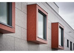 Moderne Fassade mit hervorstehenden, rotbraunen Fensterrahmen und grauen Paneelen, aufgenommen an einem bewölkten Tag.