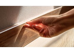 Hand drückt Holz-Sockelleiste an eine weiße Wand.