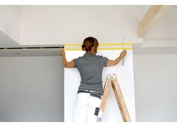 Person, die auf einer Holzleiter steht und gelbes Abdeckband entlang einer horizontalen Linie an einer weißen Wand anbringt, 