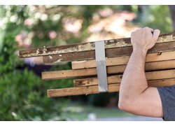 Bild zeigt eine Person, die mehrere Holzlatten, die mit silbernem Gewebeband zusammengebunden sind, auf der Schulter trägt.