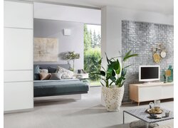 Das Bild zeigt einen hellen, modern eingerichteten Wohn- und Schlafbereich mit einem an der Wand installierten Leaf Air Lüftungsgerät. Der Raum ist offen gestaltet und kombiniert eine graue Ziegelwand mit hellen Boden- und Möbelfarben. Pflanzen, Dekorationselemente und Tageslicht aus großen Fenstern schaffen eine wohnliche Atmosphäre. Das Lüftungsgerät ist dezent in das Raumdesign integriert und sorgt für frische, gefilterte Luft.