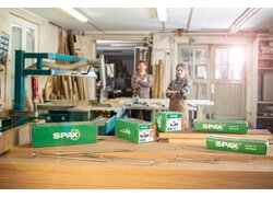 Verpackungen von SPAX Kohlenstoffstahlschrauben auf einer Werkbank, in einer Werkstatt mit zwei Handwerkern im Hintergrund