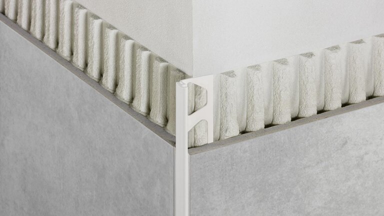 Das Bild zeigt eine Ecke, die mit einem Kantenschutzprofil in hellem Farbton versehen ist, das zwischen zwei gefliesten Flächen eingesetzt wurde, um eine saubere und geschützte Kante zu schaffen; es handelt sich um ein Produktbild für ein Kantenschutzprofil von Schlüter-Systems.