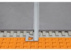 Detailansicht eines Bodenübergangs, bei dem zwei Fliesenflächen durch ein Aluminiumprofil getrennt werden. Das Profil ist eingebettet in eine Estrichunterlage mit einem orangefarbenen Entkopplungselement 