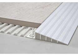 Höhenübergang zwischen einer gefliesten Oberfläche und einer tiefer liegenden Betonfläche, der durch ein rampenförmiges Aluminiumprofil ausgeglichen wird.