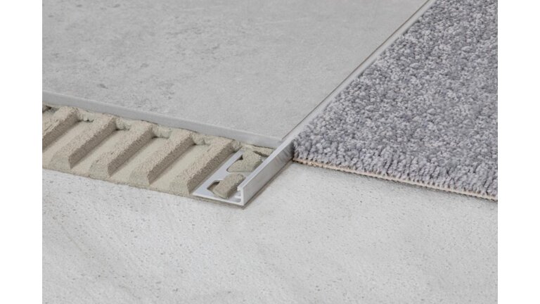  Nahaufnahme der Verbindung zwischen einem grauen Teppich und einem Betonboden. Ein Aluminiumprofil, das sichtbar in die Estrichunterlage eingebettet ist, trennt und stabilisiert die beiden Bodenbeläge.