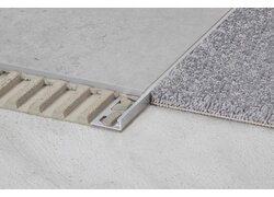  Nahaufnahme der Verbindung zwischen einem grauen Teppich und einem Betonboden. Ein Aluminiumprofil, das sichtbar in die Estrichunterlage eingebettet ist, trennt und stabilisiert die beiden Bodenbeläge.