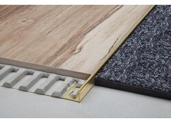 Ein Messingprofil bildet die präzise und saubere Verbindung zwischen einer Holzoptik-Laminatfläche und einem grauem Teppich.