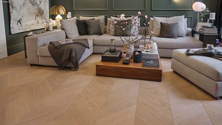 mafi Naturholzboden Eiche in einem Wohnzimmer. man sieht ein großes Sofa, davor ein Couchtisch und dahinter eine mit Holz verkleidete Wand in grün. Auf dem Tisch stehen Magnolienäste mit Blüten