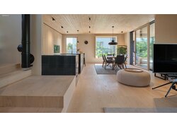 mafi Naturholzboden Esche in einem Wohn- Essbereich mit offener Küche. Auch die Wand ist Holzverkleidet. Der Raum ist durch große Fenster und eine Terrassentür einladend hell.