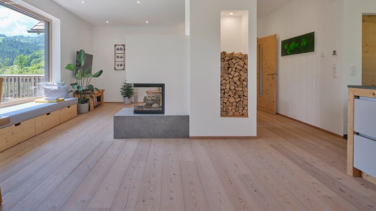 Heller mafi Naturholzboden aus Nadelholz in einem Wohnraum. Mittig ist ein Kamin und links eine Fensterreihe mit Aussicht in die Berge
