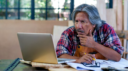 Alleinstellungsmerkmal Alter asiatischer Handwerker oder Tischler schaut auf den Bildschirm des Laptops und denkt über seinen Arbeitsprozess nach und sitzt in der Nähe von Papier- und Holzprodukten am Arbeitsplatz.