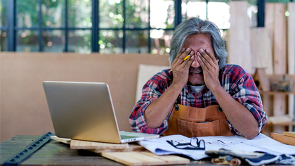 Krankheit Alter asiatischer Handwerker oder Tischler schaut auf den Bildschirm des Laptops und denkt über seinen Arbeitsprozess nach und sitzt in der Nähe von Papier- und Holzprodukten am Arbeitsplatz.